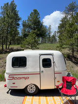 Enjoying nature with an RV rental: a white Scamp trailer by pine trees with a red chair and striped mat for outdoor relaxation.