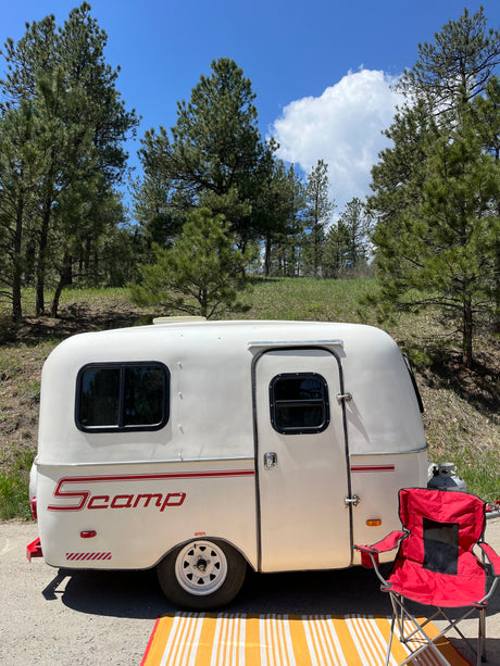 Enjoying nature with an RV rental: a white Scamp trailer by pine trees with a red chair and striped mat for outdoor relaxation.