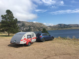 A blue Subaru tows a grey teardrop RV rental, parked by a scenic lake and mountains for camping/travel.