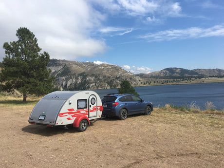A blue Subaru tows a grey teardrop RV rental, parked by a scenic lake and mountains for camping/travel.