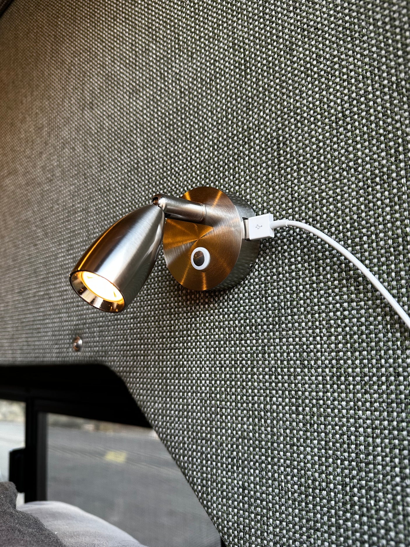 Close-up of a brushed metal reading light and USB port, mounted on a green woven fabric wall, ideal for an RV rental. Light glows warmly.