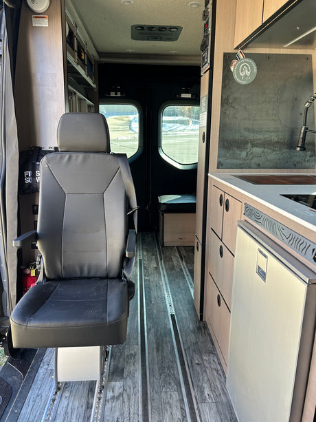 View inside an RV rental showing a front swivel seat, compact kitchen, and floor tracks for adaptable living.