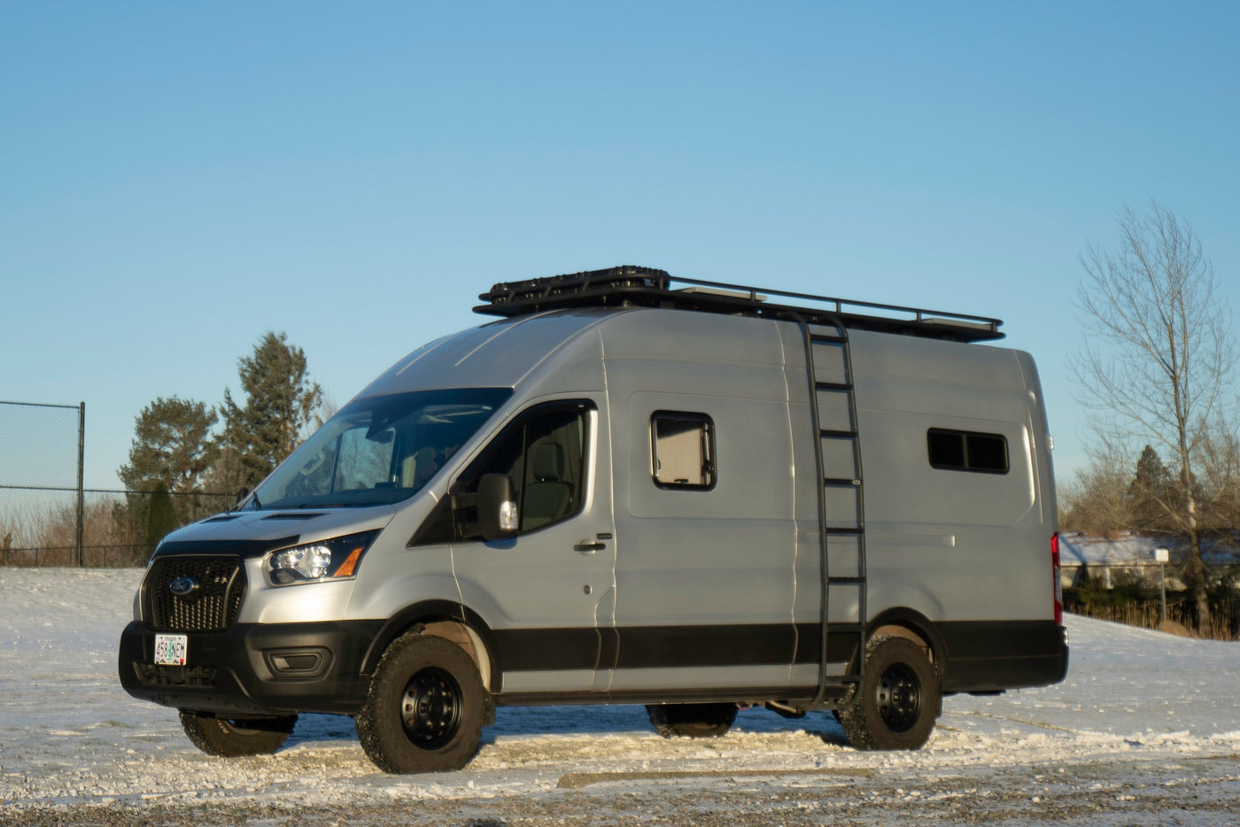 Silver Ford Transit camper van, ideal for RV rental, with roof rack and ladder on snowy terrain under clear sky. — photo 25