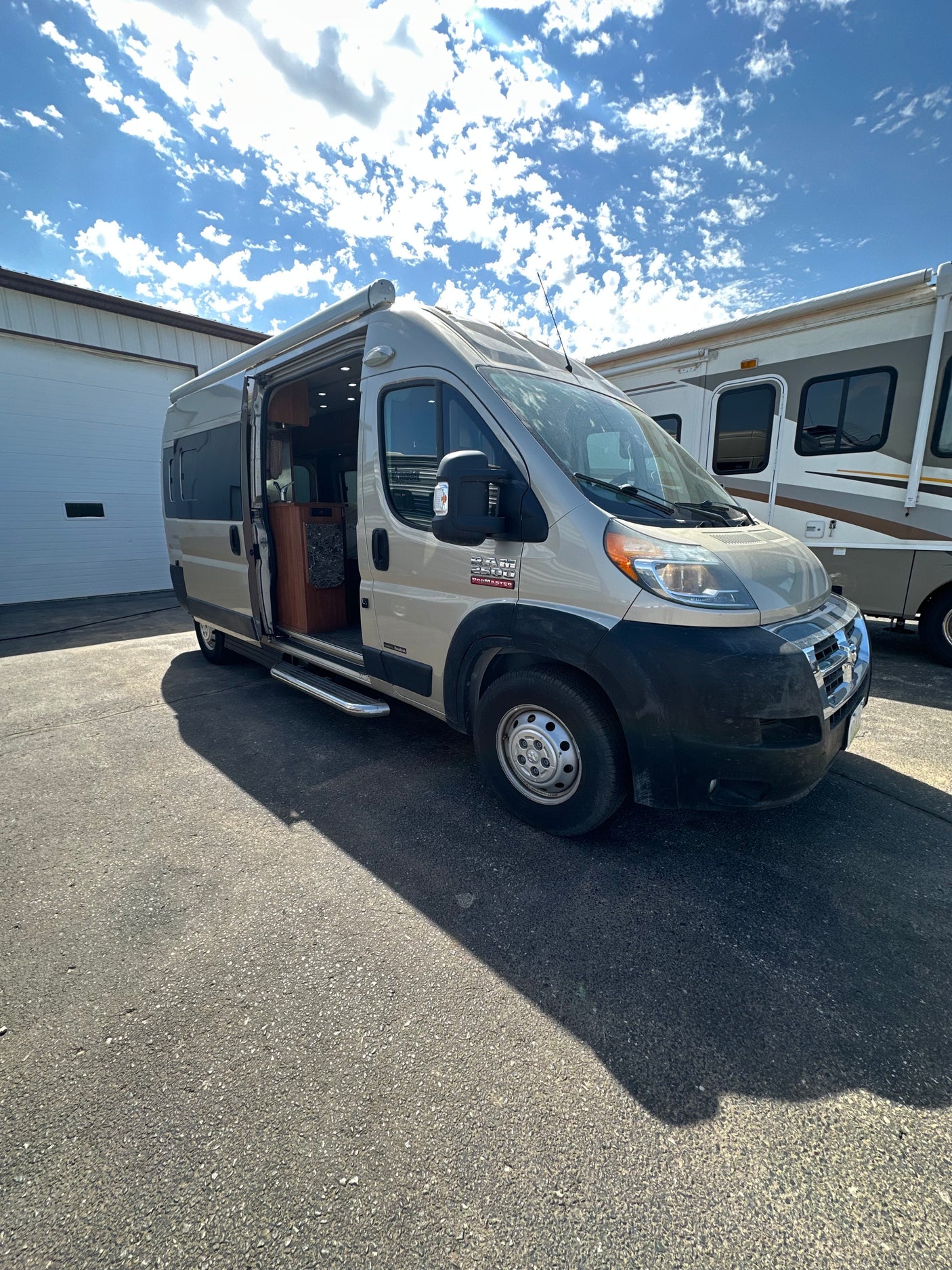 A tan Ram ProMaster camper van with an open door, revealing its interior, is parked for rv rental viewing. Another large RV is nearby.