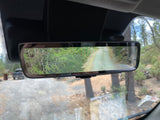 An RV rental's digital rearview mirror displays a rear camera view of a wooded path, enhancing driving visibility.