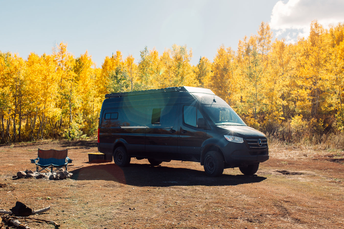 Black Mercedes Sprinter camper van (RV rental) parked among golden autumn aspens, with a blue camp chair by a fire pit.