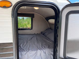 Cozy rv rental interior: a comfy bed with grey sheets, ceiling light, and window revealing green trees outside.