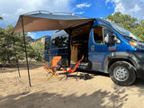 Enjoy a spacious RV rental setup: blue camper van with extended awning, camping chairs, and scenic mountain views.