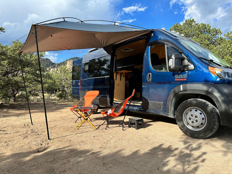 Enjoy a spacious RV rental setup: blue camper van with extended awning, camping chairs, and scenic mountain views.