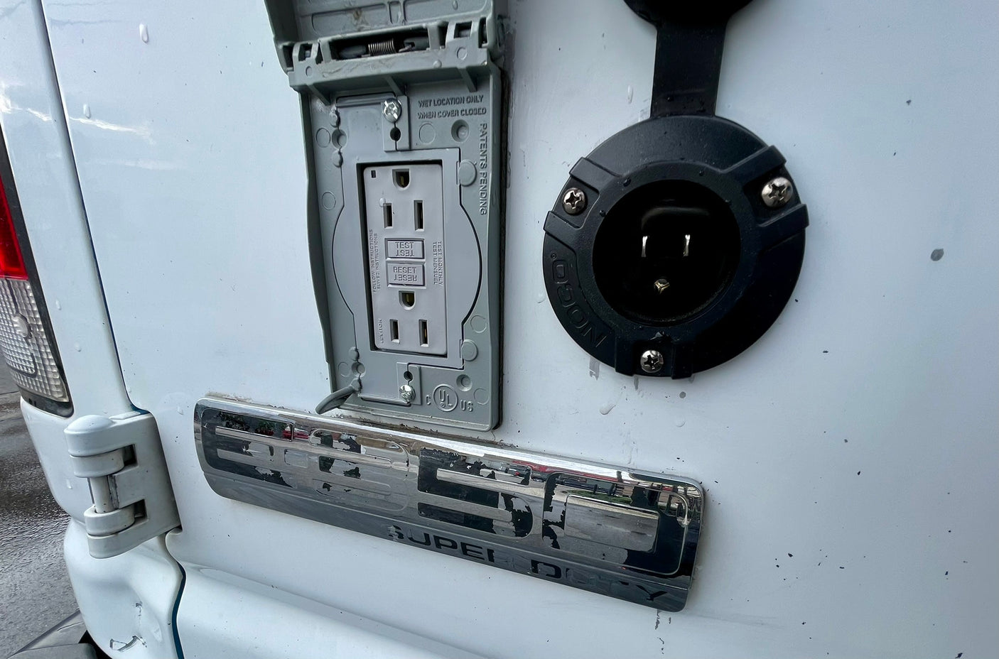 RV rental: Exterior of a white utility van featuring a weatherproof GFCI outlet and a black shore power inlet for outdoor electrical access.