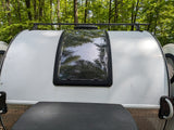White teardrop RV rental parked in a forest. Features a roof rack, large window reflecting green trees, and lower textured trim.