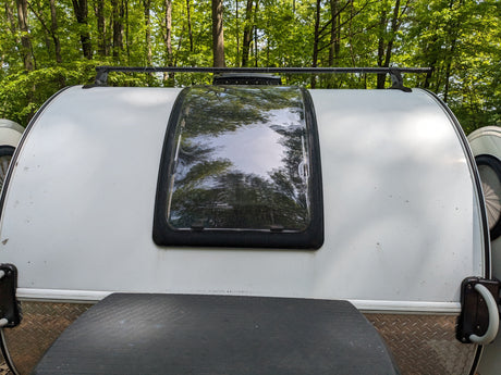 White teardrop RV rental parked in a forest. Features a roof rack, large window reflecting green trees, and lower textured trim.
