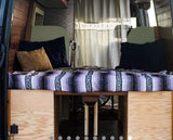 RV rental van interior: A cozy bed with a striped blanket & pillows, flanked by wood walls. Under-bed storage visible. White curtains in back.