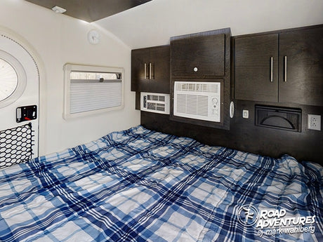 Cozy rv rental bedroom interior with a plaid bed, overhead cabinets, window, and AC units for comfortable travel.