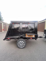 This black, custom-built camping trailer, ideal for unique rv rental experiences, features a window and outdoor shelf.