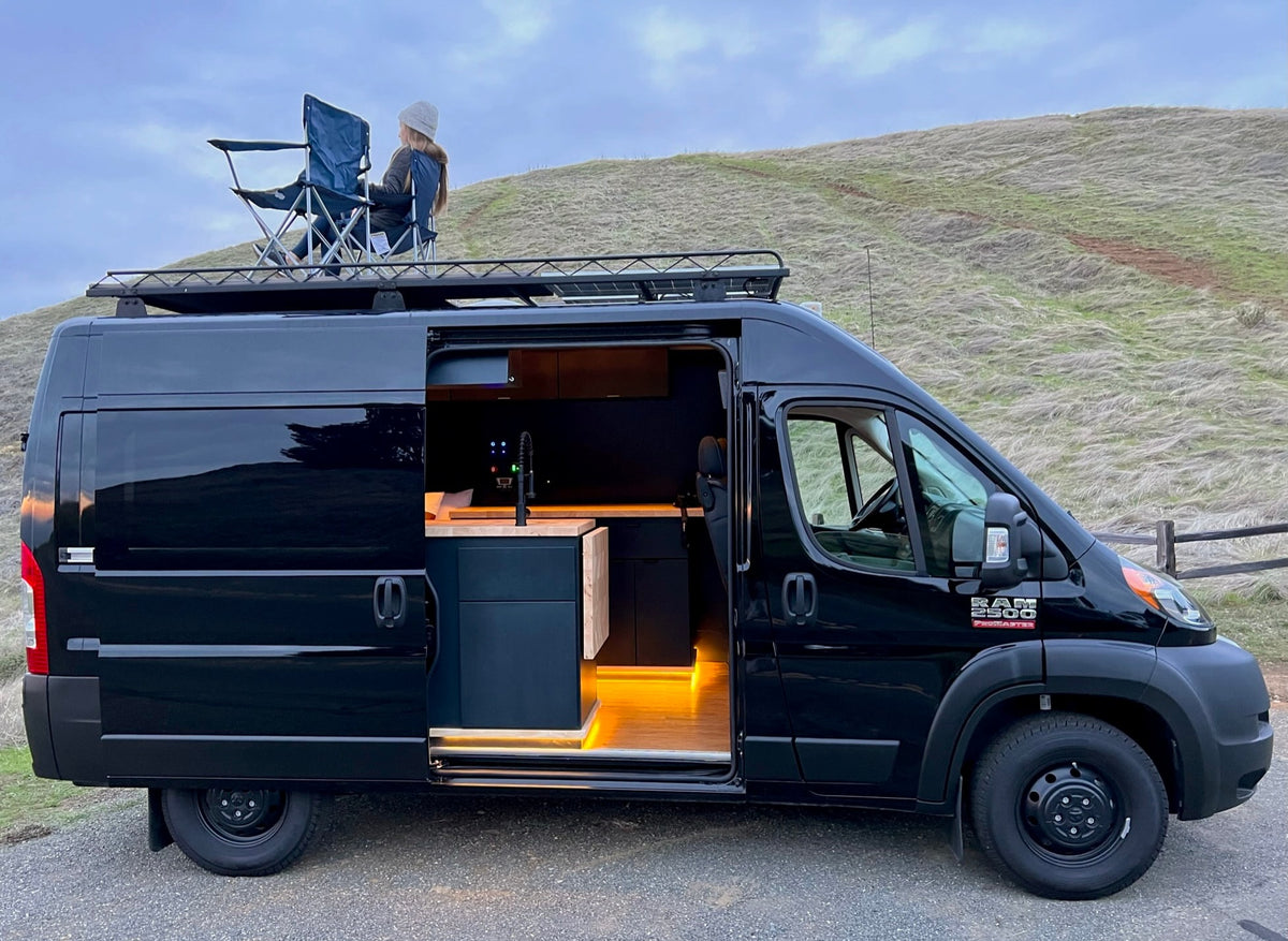 This black RV rental camper van shows its stylish interior & a person relaxing on the roof rack, ready for adventure. — photo 2