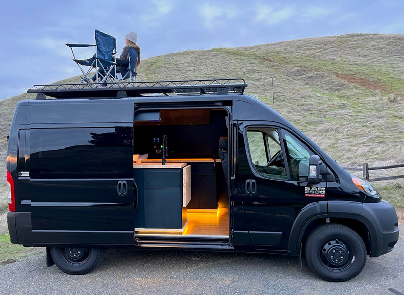 This black RV rental camper van shows its stylish interior & a person relaxing on the roof rack, ready for adventure. — photo 2