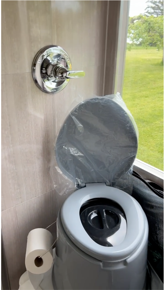 Compact RV rental bathroom featuring a portable toilet, shower valve, and nature view outside. Toilet paper handy. — photo 7