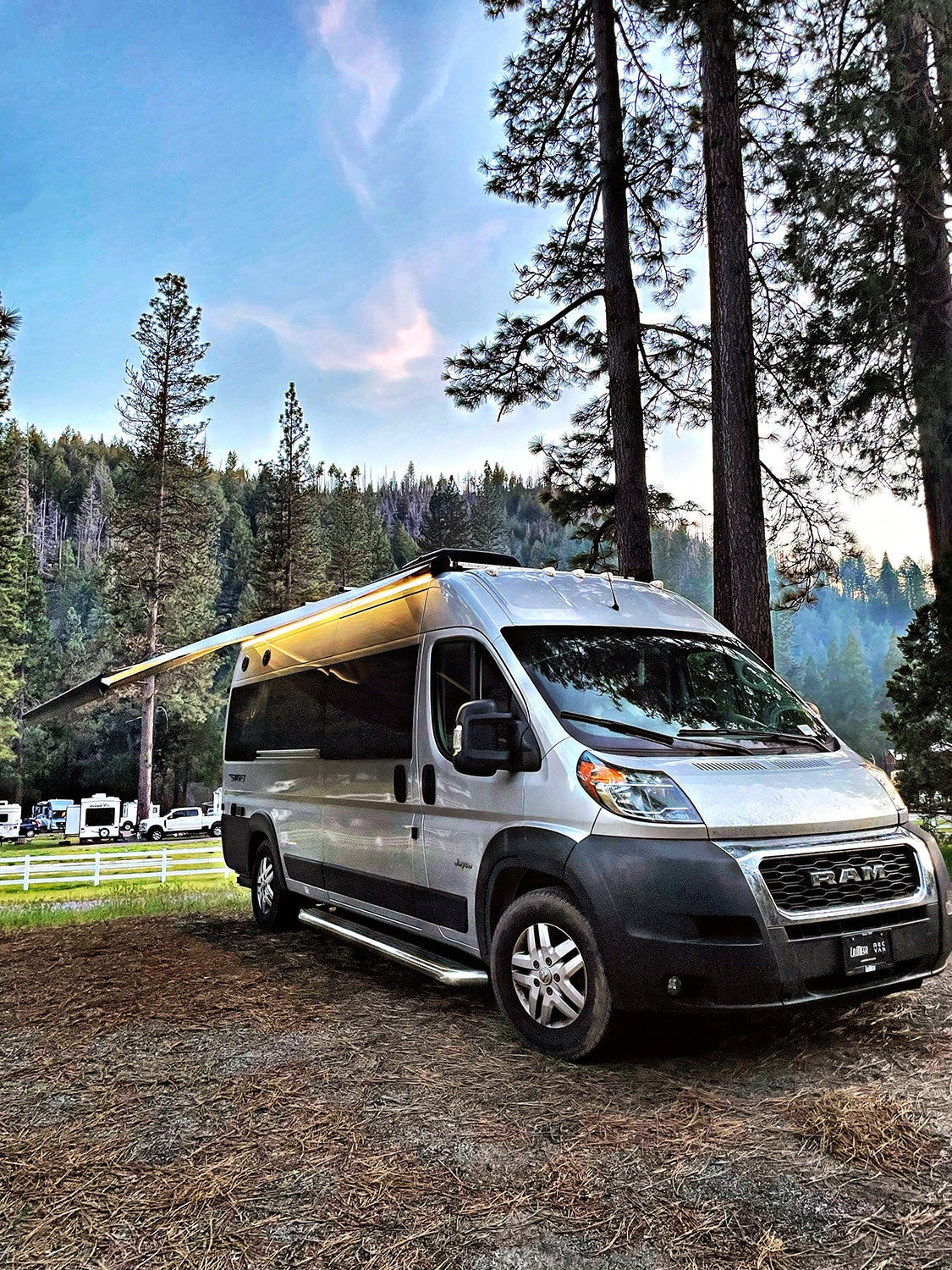 Silver camper van RV rental with awning extended, parked in a forest campground, ready for your next outdoor escape.