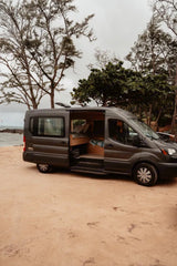 RV rental: A dark grey camper van parked on a sandy beach. Its open door reveals a cozy wooden interior, perfect for coastal travel.