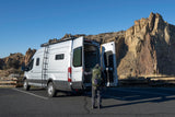 Adventure beckons! A person by an open silver Transit camper van at rocky cliffs. Ideal rv rental for exploration.