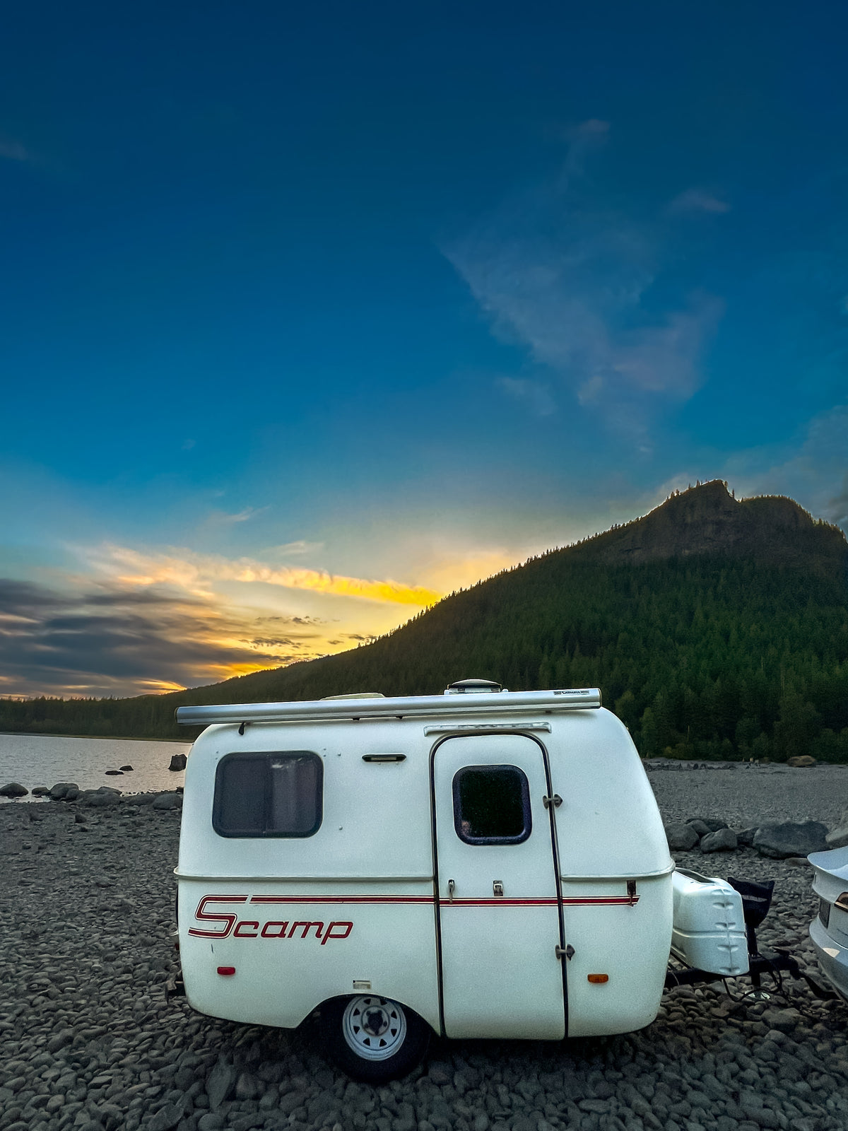 Experience the beauty of nature with an RV rental! This white Scamp camper sits by a lake at sunset, framed by a forested mountain. — photo 6