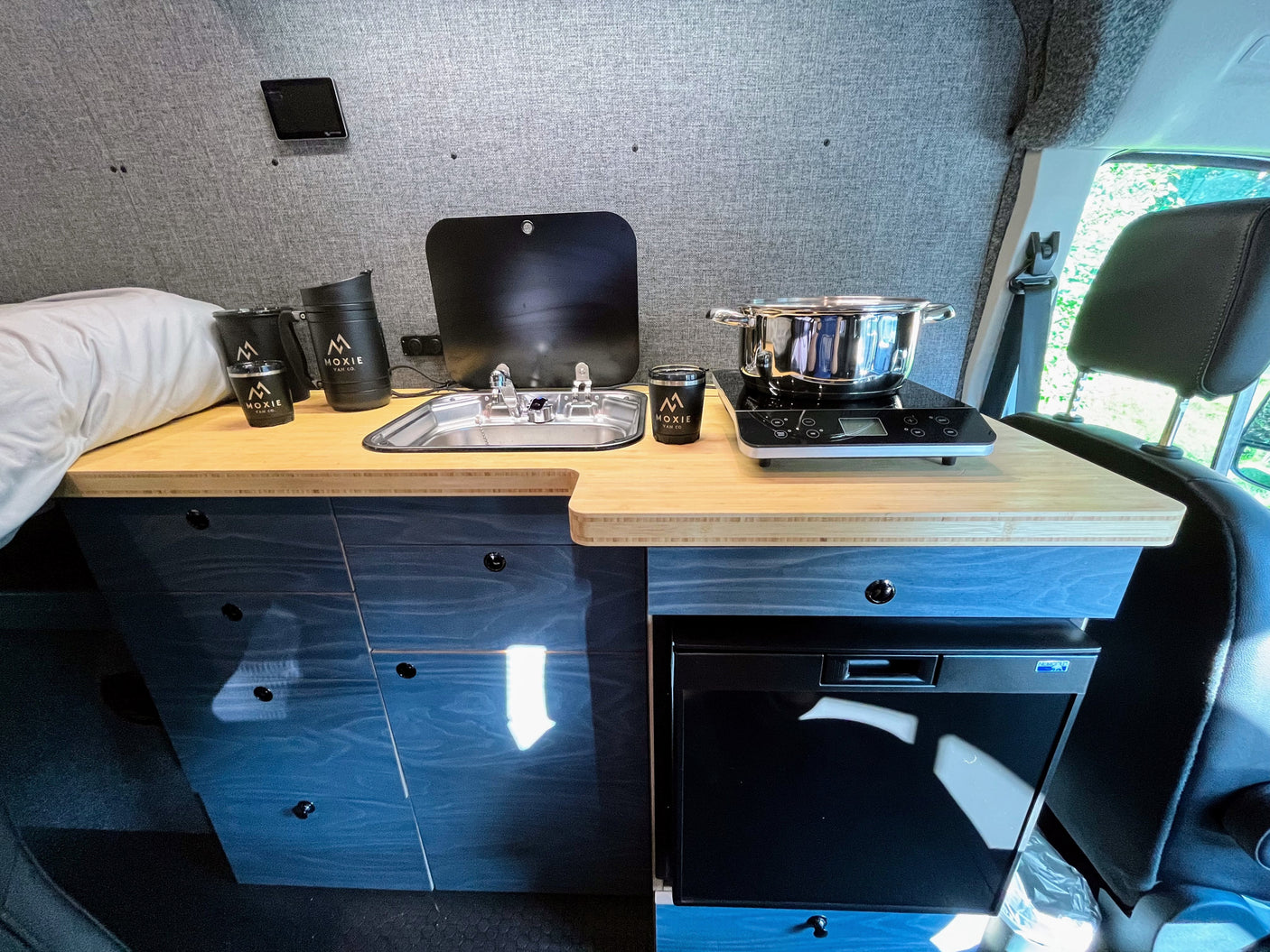 Moxie Van Co. RV rental: a compact camper van kitchen with a sink, induction cooktop, and under-counter refrigerator.