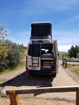 White pop-top camper van with open rear doors on a dirt path in nature. Explore with an RV rental for your next adventure!