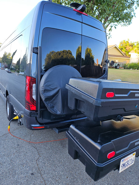Rear of a dark RV rental van, showcasing dual storage boxes, a spare tire, and an orange power cable plugged in.