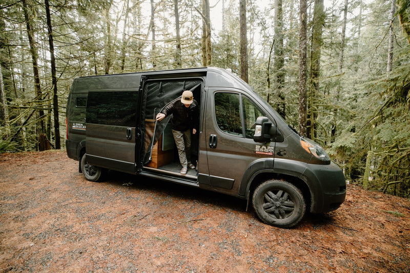 A person in a cap and jacket steps from a grey RV rental van, parked amidst moss-covered trees on a forest path. Ideal for outdoor adventures. — photo 11