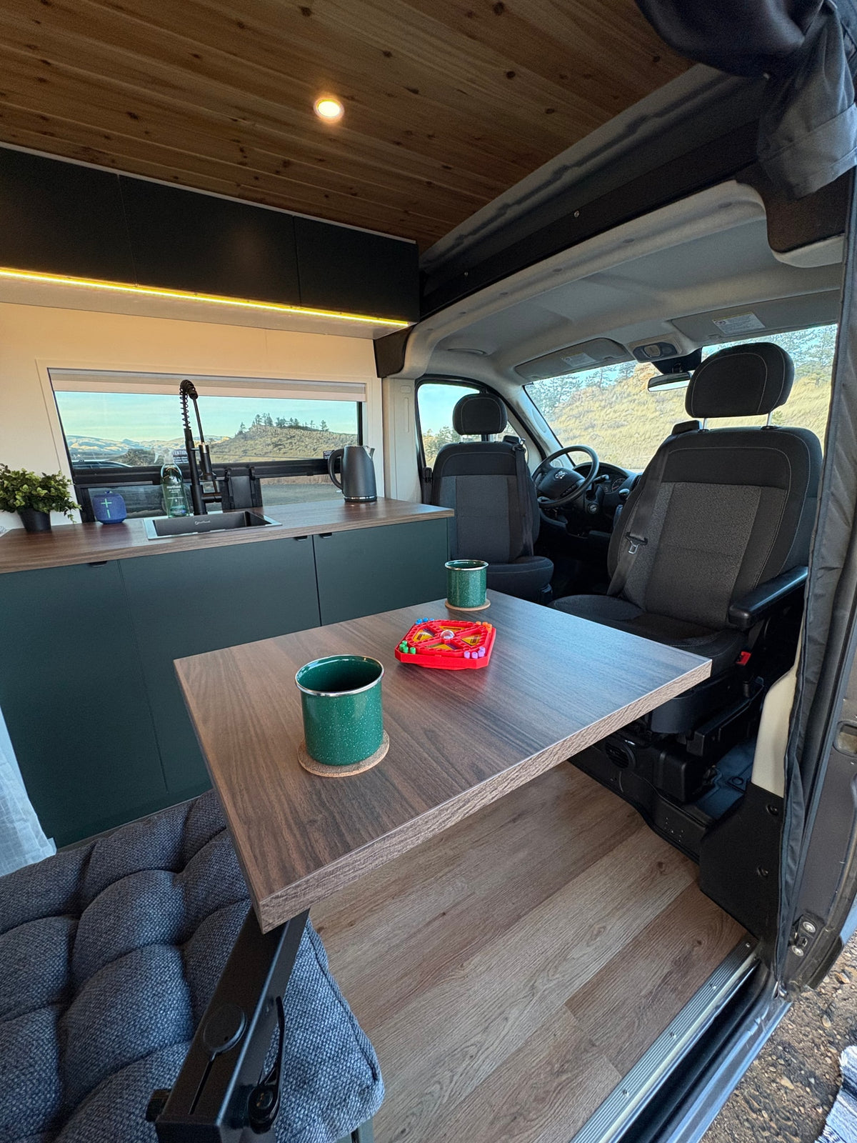 Modern RV rental interior: kitchen with sink, dining table with mugs & game, comfy seating, and scenic window view. — photo 9