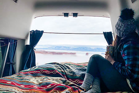  and front seating in a camper van.|RV rental interior: a compact sink with blue water tanks for fresh and greywater stored below. A bed is adjacent.|RV rental adventure prep: A woman with a red bandana and tattoos stocks her cooler with a thermos — photo 6
