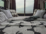 Cozy RV rental bed with patterned pillows & moose blanket, offering a beautiful beach view through the back screen.
