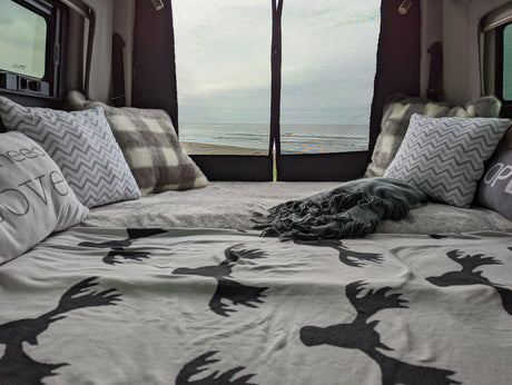Cozy RV rental bed with patterned pillows & moose blanket, offering a beautiful beach view through the back screen.