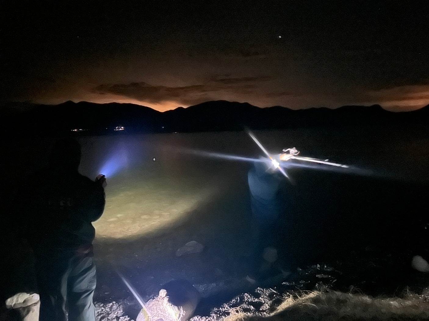 Exploring the tranquil lake at night, lit by flashlights, a perfect end to an RV rental adventure. Mountains loom under starry skies.