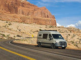A light grey RV rental van on a desert road, with towering red rock formations in the background. Perfect for scenic road trips.
