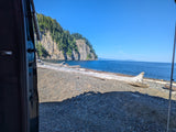 View from an RV rental: Rocky beach with driftwood, calm blue sea, and a tree-covered cliff.
