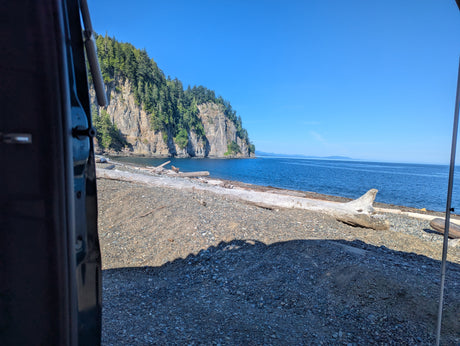 View from an RV rental: Rocky beach with driftwood, calm blue sea, and a tree-covered cliff.