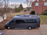 Dark grey RV rental camper van with solar panels & AC on its roof, parked on a residential driveway.