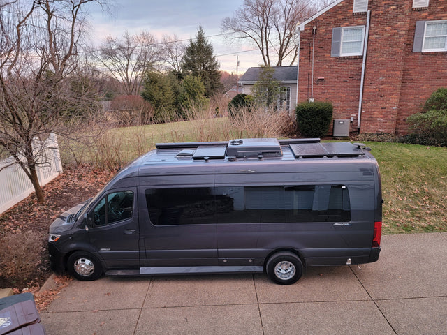 Dark grey RV rental camper van with solar panels & AC on its roof, parked on a residential driveway.
