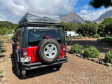 Red Jeep RV rental with rooftop tent, parked at a Maui campsite. Mountains and trees in the background, perfect for adventure.