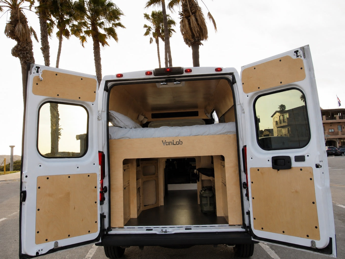 RV rental: Custom camper van interior with bed and storage, seen from open rear doors. Palm trees and coast outside.