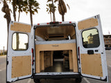 RV rental: Custom camper van interior with bed and storage, seen from open rear doors. Palm trees and coast outside.