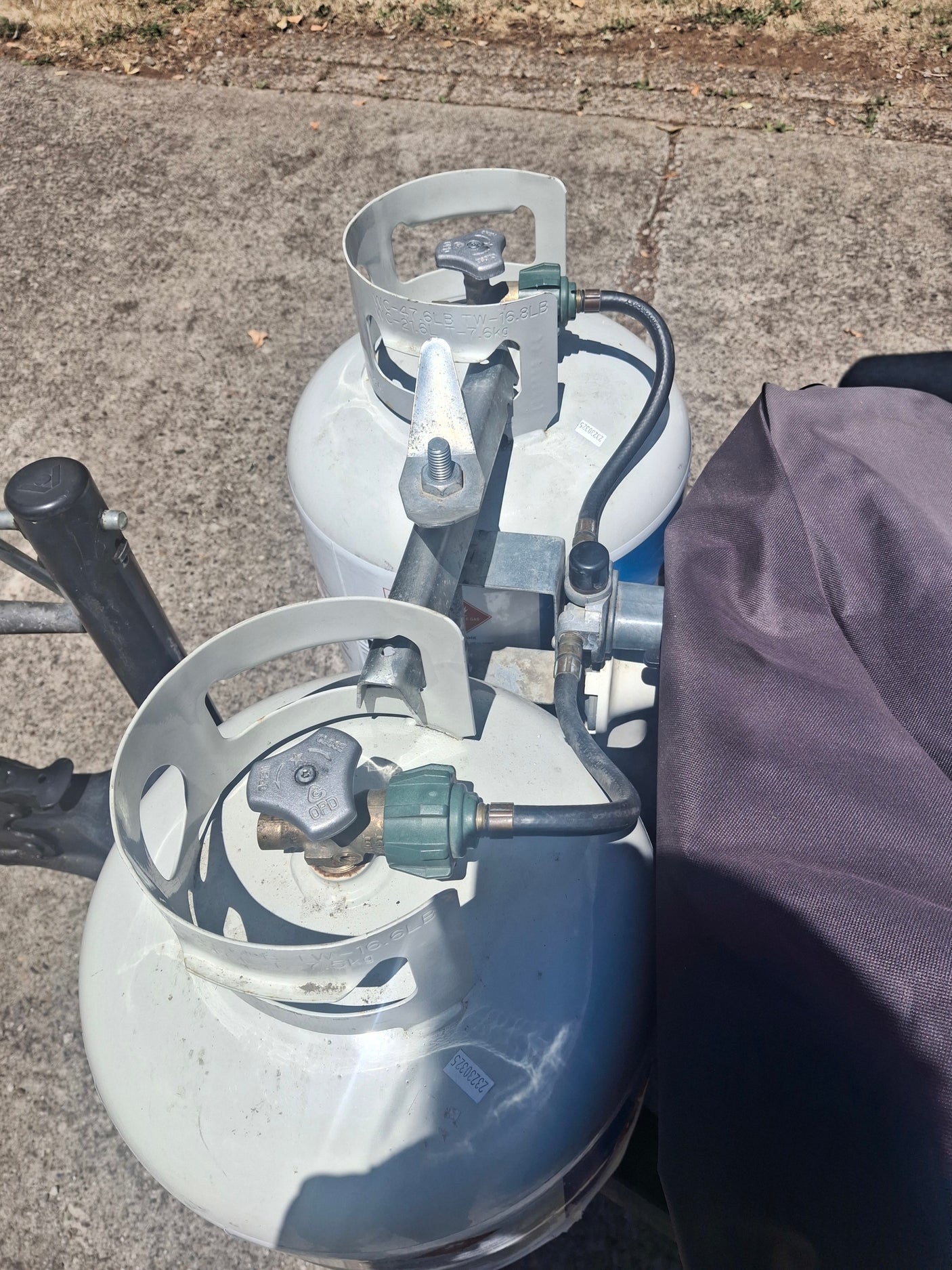 Two white propane tanks, essential for an RV rental, are connected by hoses and a regulator, with a dark cover nearby.