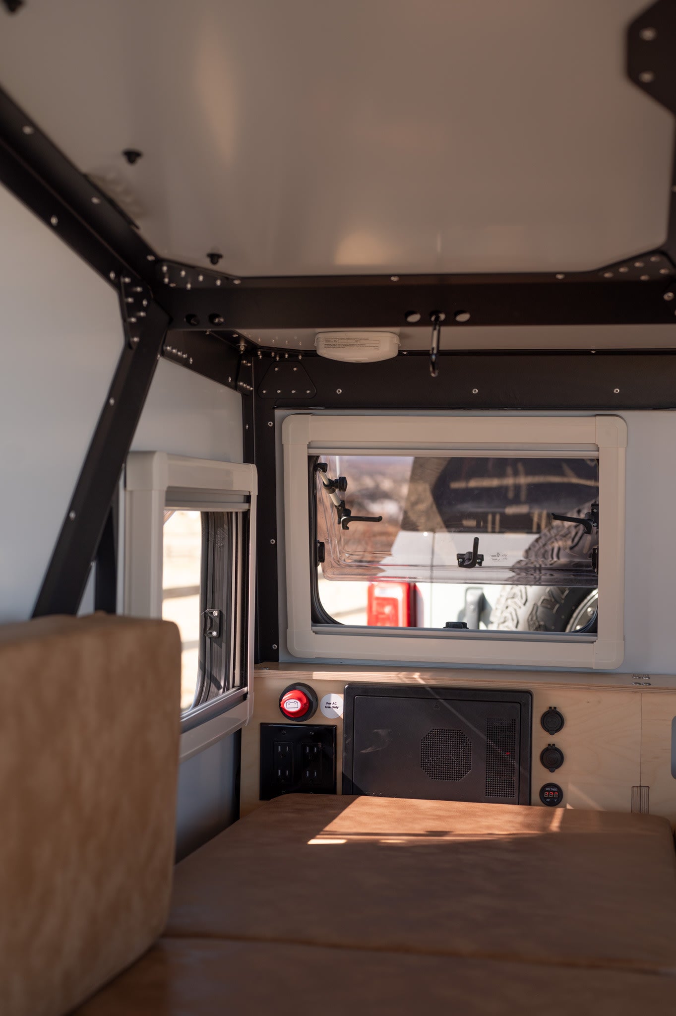 Interior of an RV rental showing a bed, open windows, and control panel with outlets. A compact, functional space.