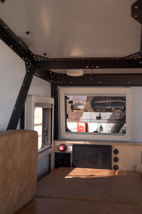 Interior of an RV rental showing a bed, open windows, and control panel with outlets. A compact, functional space.