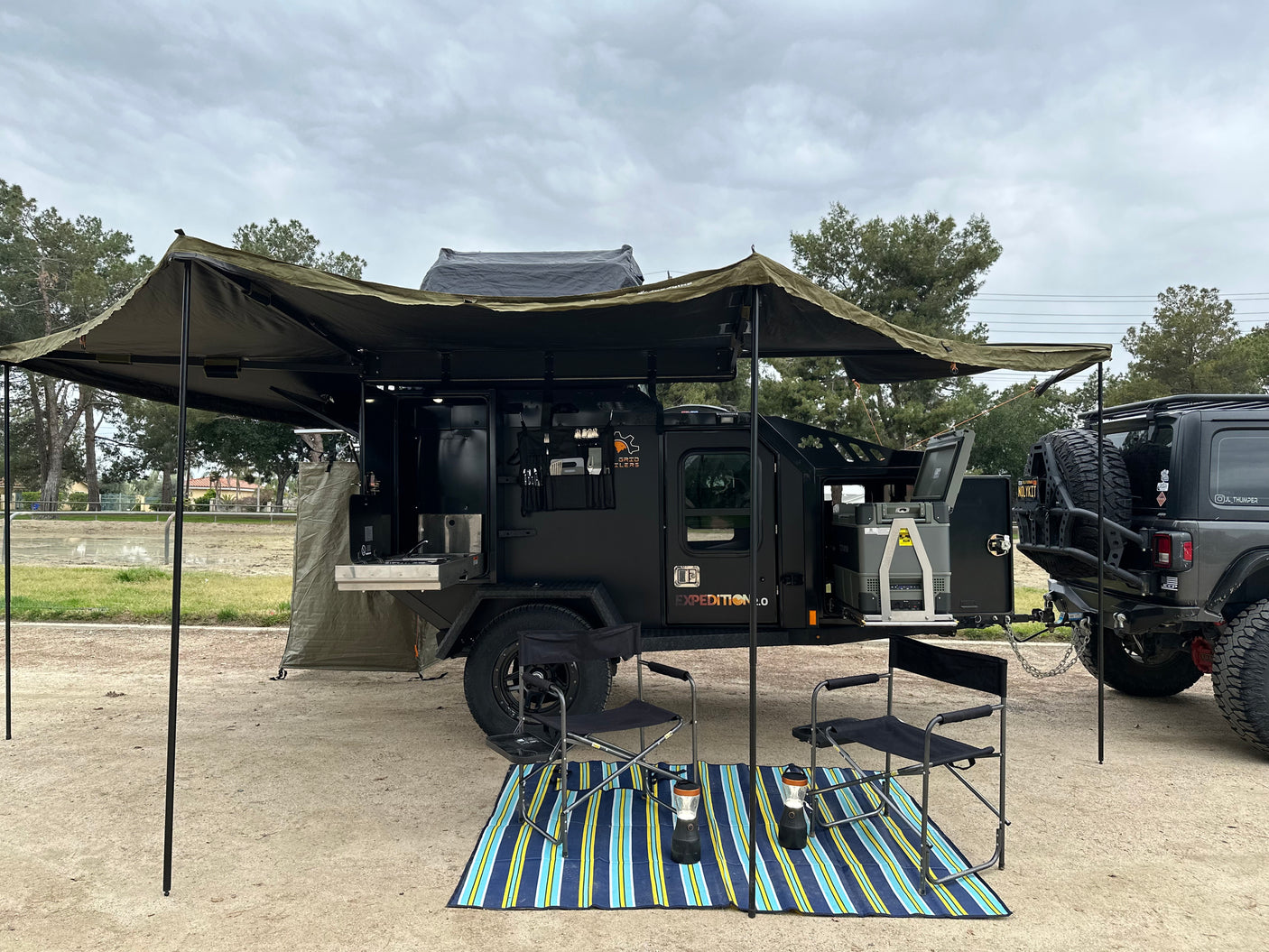 This rugged overlanding trailer, ideal for rv rental, offers an outdoor kitchen, fridge, and large awning, hitched to an off-road Jeep.