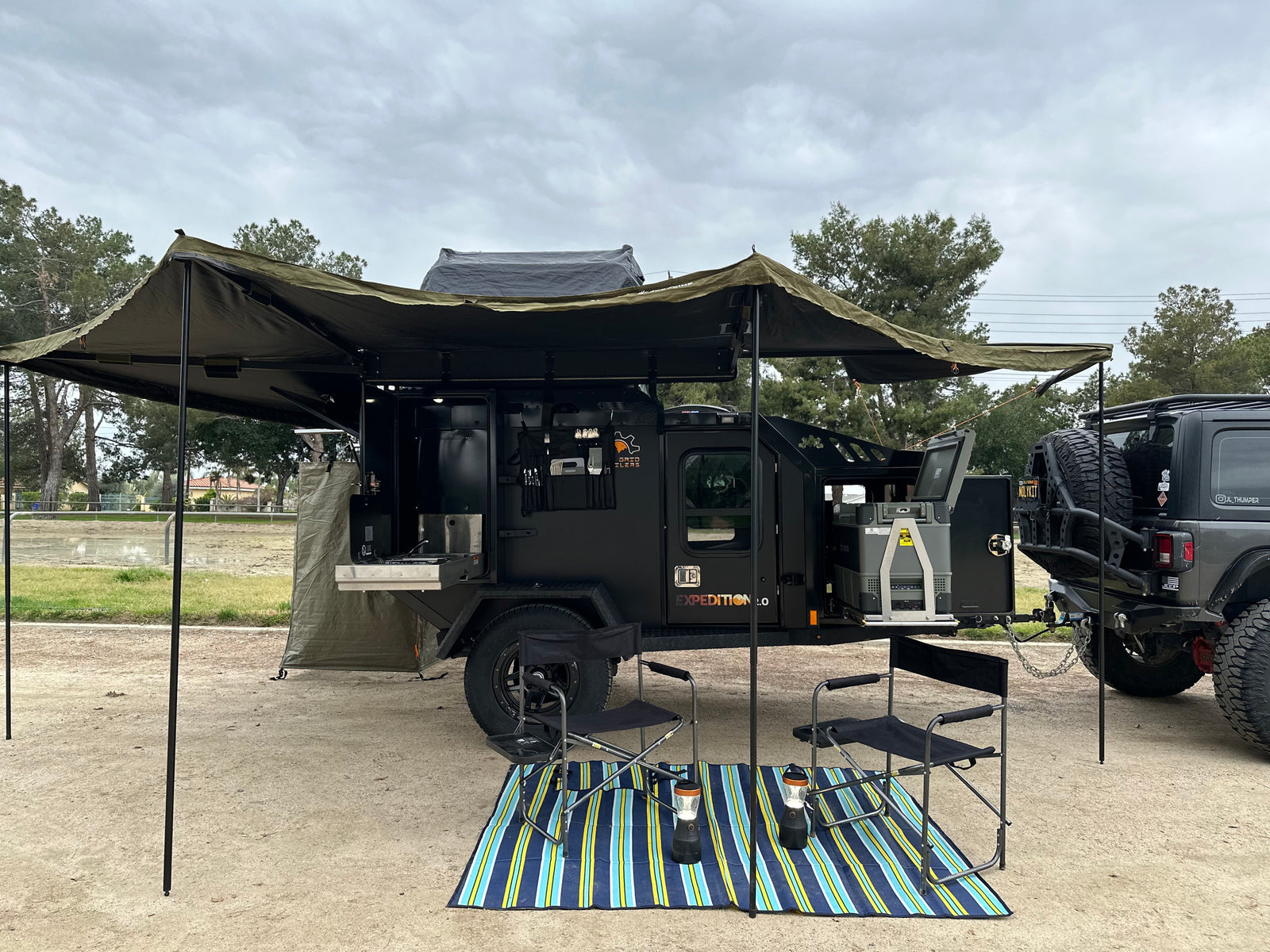 This rugged overlanding trailer, ideal for rv rental, offers an outdoor kitchen, fridge, and large awning, hitched to an off-road Jeep.
