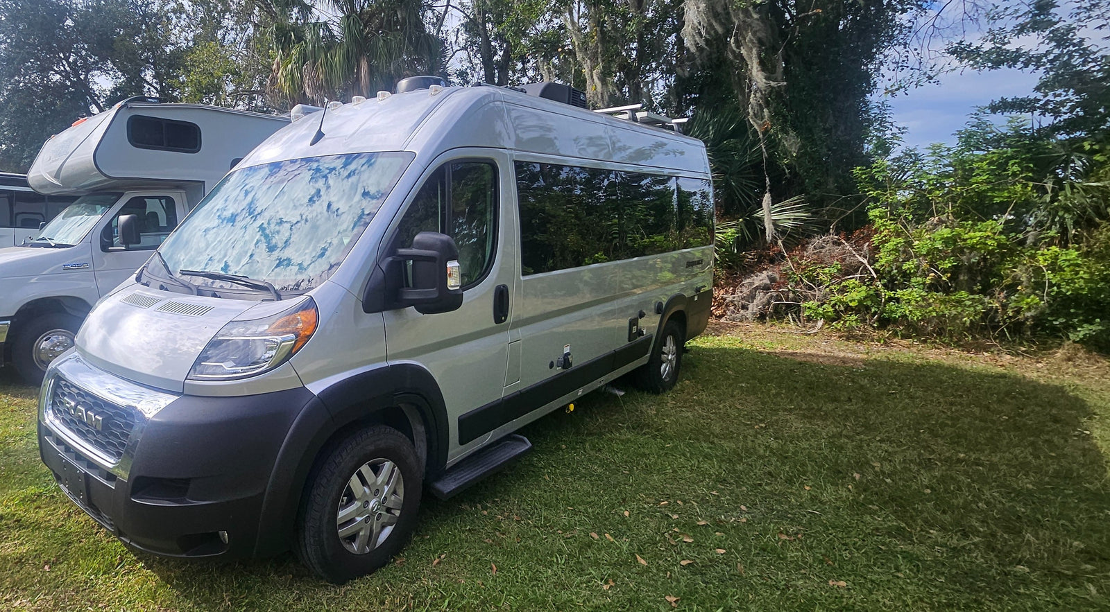 Ready for adventure! This silver Ram Promaster camper van RV rental sits on green grass, backed by trees, with another RV nearby.