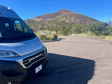 Silver Winnebago RV rental van on a winding road, facing a volcanic mountain with sparse trees under a blue sky. Great for travel!